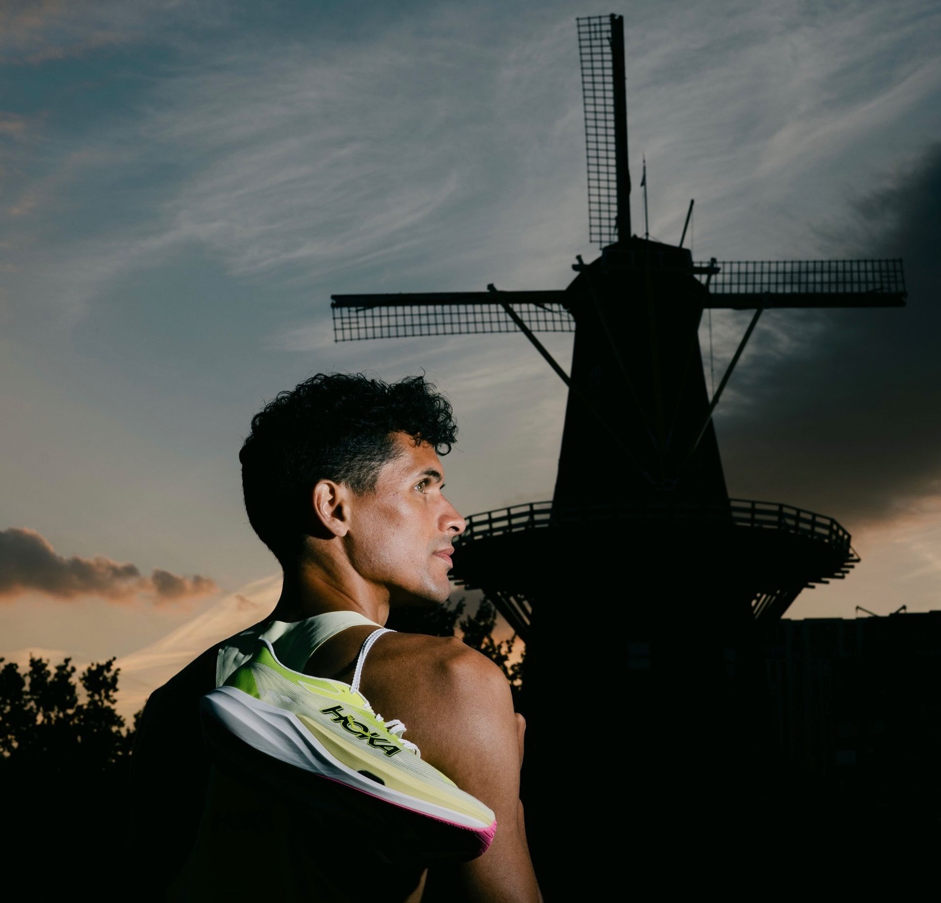 Noah at dusk with a HOKA racing shoe on his shoulder, a Dutch windmill behind him